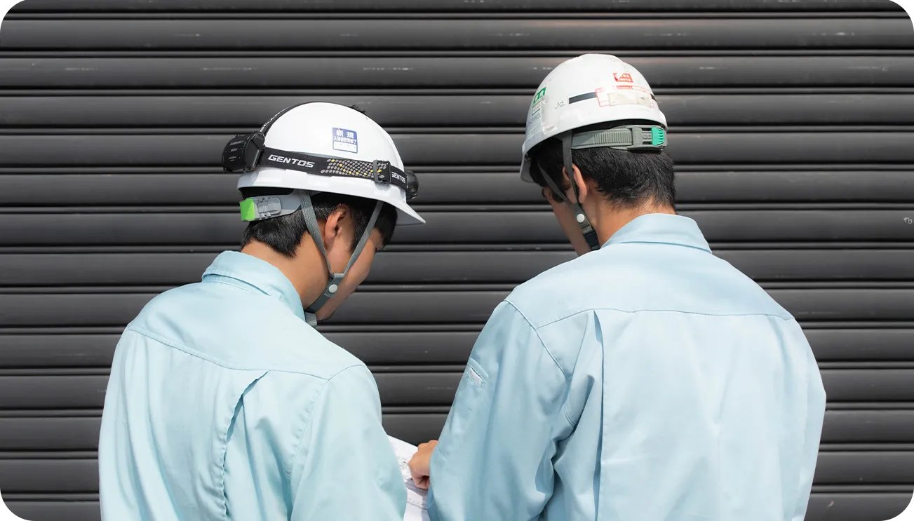 設計図を確認する作業服の男性2人の後ろ姿